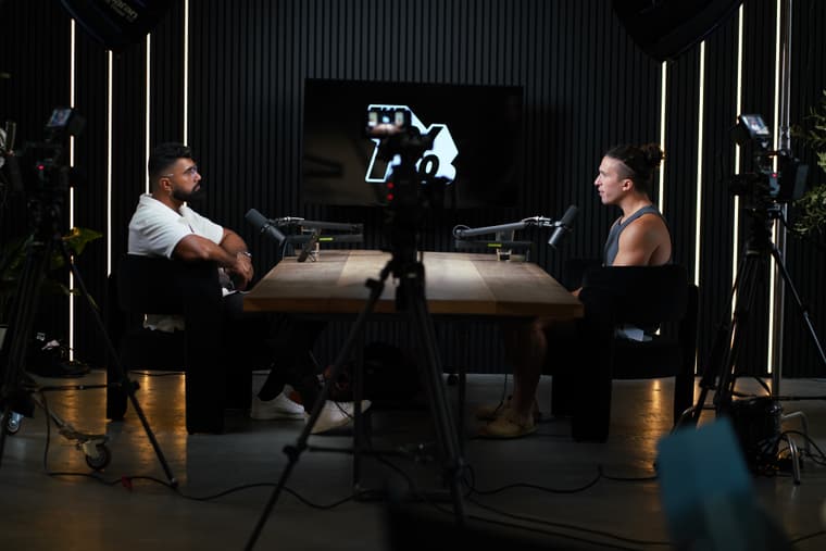 Deepak and a guest face each other across the table on The 1% Show set, with a camera in the foreground.